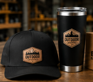 A black trucker cap and a matching black insulated tumbler with stainless steel trim, both printed with a brown outdoor lifestyle shield logo, displayed on a wooden table with a blurred rustic background.
